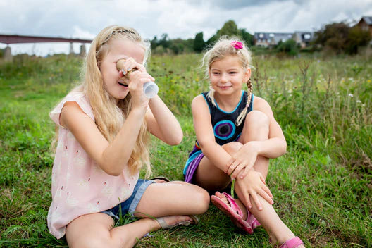 Two young girls sit on the grass; one looks through a Huckleberry Kaleidoscope while the other, a young artisan, carves with a kid-friendly wood carving knife. They are in an open field with cloudy skies in the background.