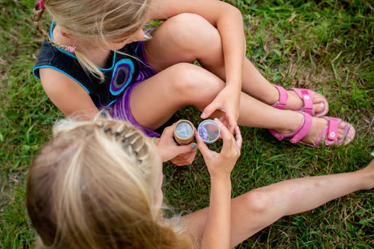 Two young artisans sit on the grass, closely examining small Huckleberry Kaleidoscopes. One child wears pink sandals while the other sports a braid and a colorful outfit.