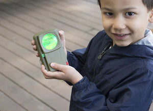 Young boy showing a Huckleberry Morse Code Light with a green glowing screen, smiling, outdoors on a wooden deck.