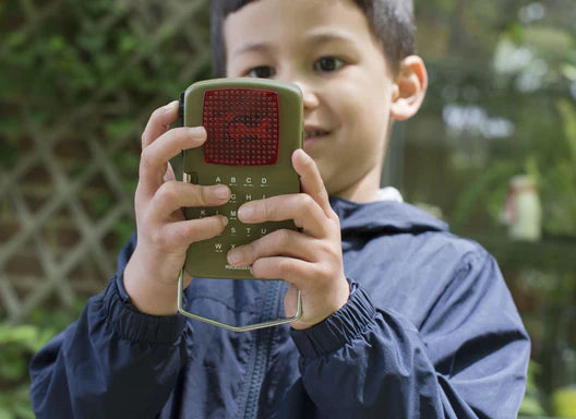 Young boy holding a red Huckleberry Morse Code Light outdoors, partially obscuring his face, with a garden background.