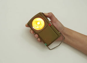 A hand holding a small, olive green Huckleberry Morse Code Light with a glowing yellow light, against a plain light background.