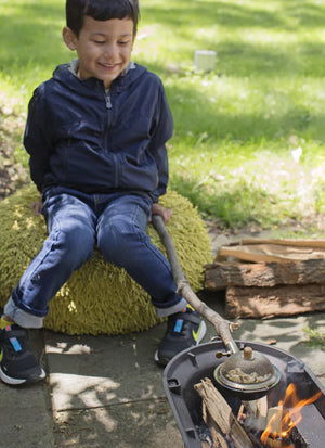 A young child in a dark jacket and jeans sits on a cushioned seat, smiling and holding a Huckleberry Popcorn Maker over a small fire pit outdoors, enjoying some campfire cooking.