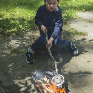 A child sits outdoors holding a Huckleberry Popcorn Maker over a small campfire, surrounded by greenery and pavement.