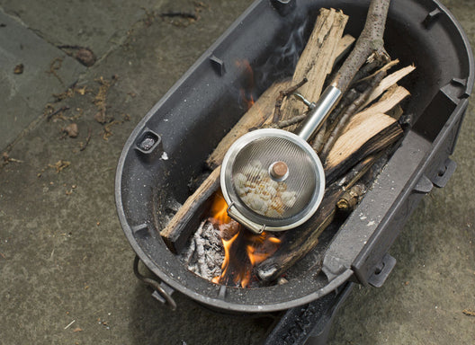 The Huckleberry Popcorn Maker is a metal container designed for campfire cooking, featuring a compartment for burning wood and a section to pop popcorn over the flames, which comes with partially popped kernels.