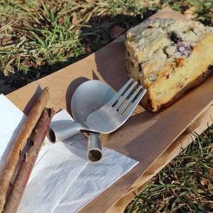 A slice of berry loaf cake on a paper plate with Huckleberry Forest Cutlery and cinnamon sticks on grass.