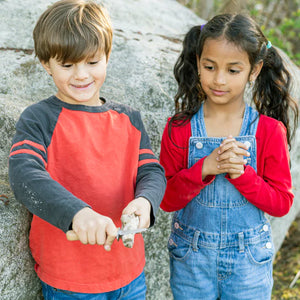 Two children stand outdoors. The boy, wearing a red and black shirt, uses the Huckleberry Carving Knife on a rock, showcasing his practical life skills. The girl in red and denim overalls watches attentively. Both kids are smiling, their creativity shining through as they explore together.