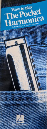 A pocket harmonica in C peeks out of denim jeans beneath the book "How to Play Pocket Harmonica.
