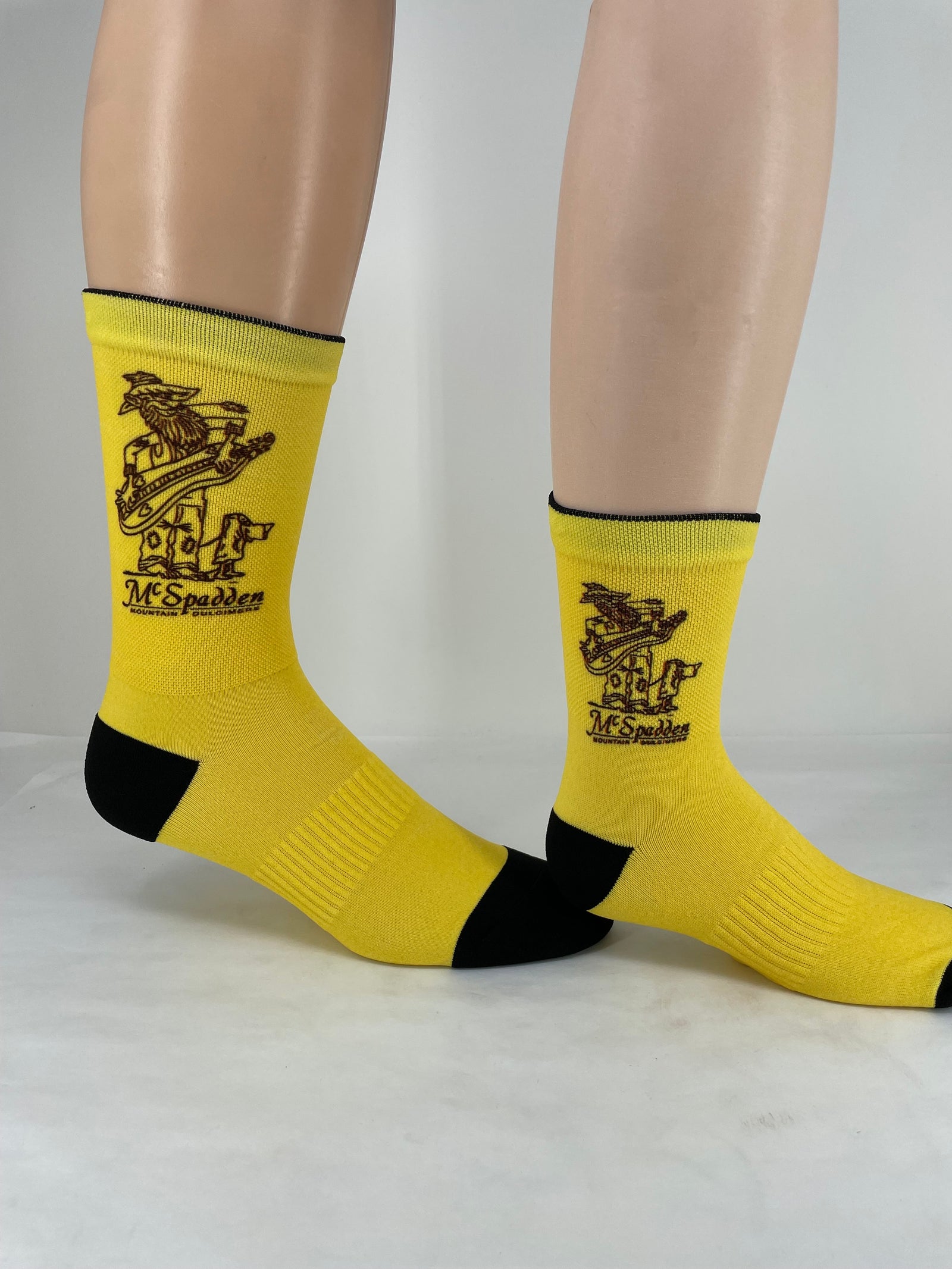 A person wearing Socks-McSpadden Hillbilly—a yellow pair with black toes and heels, graphic design, and "McSpadden" text on the sides—stands on a white surface.