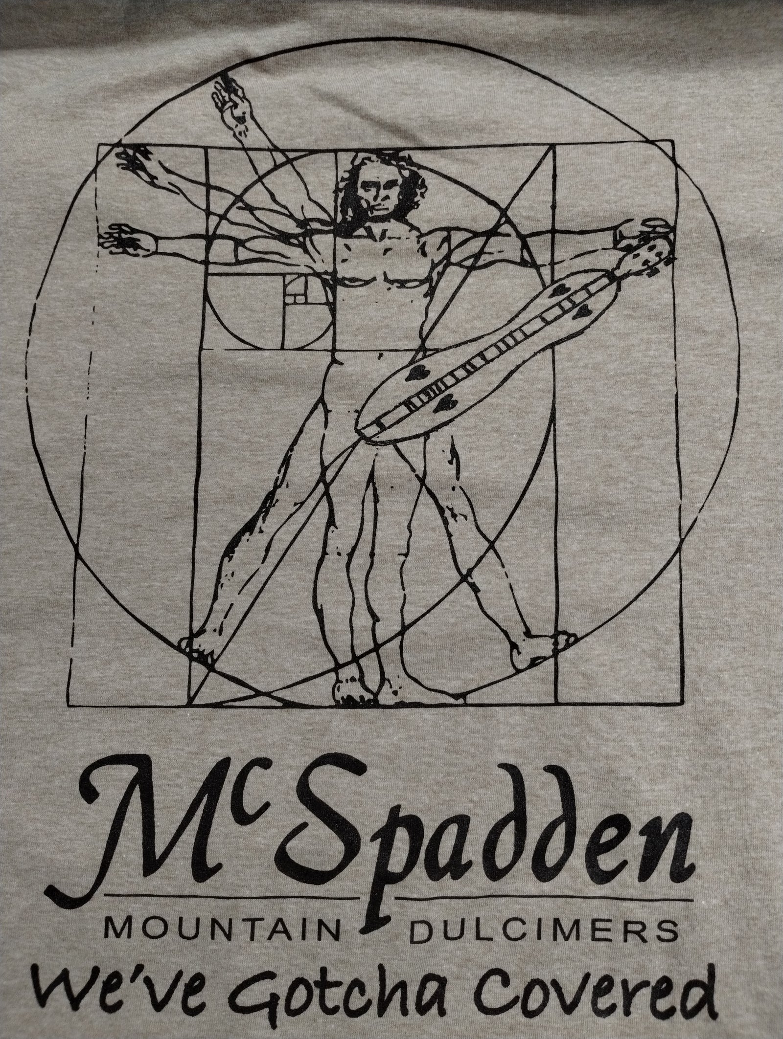 Tan shirt featuring a line drawing of the Vitruvian Man holding a dulcimer, with “We’ve Gotcha Covered” text and the McSpadden Mountain Dulcimers logo; product name: Shirt with Vitruvian Man (We've Gotcha Covered).