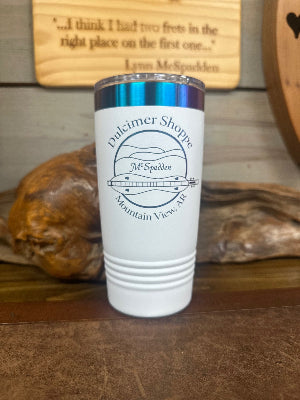 The McSpadden Dulcimer Shoppe Insulated Coffee Mug, featuring “Dulcimer Shoppe Mountain View, AR” and a dulcimer logo, sits on a wooden surface with decorative items in the background.