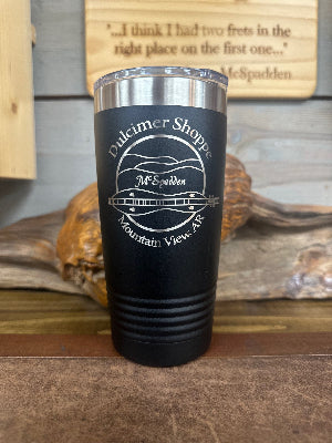 The McSpadden Dulcimer Shoppe Insulated Coffee Mug is a sleek black mug featuring “Dulcimer Shoppe Mountain View, AR” and a dulcimer graphic printed in white. Resting on a wooden surface, this insulated mug is perfect for keeping your beverages hot or cold while you’re on the go.