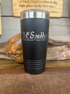 The McSpadden Dulcimer Shoppe Insulated Coffee Mug, featuring “McSpadden Mountain Dulcimers” printed on its sleek black exterior, is displayed on a wooden surface with rustic decor in the background.