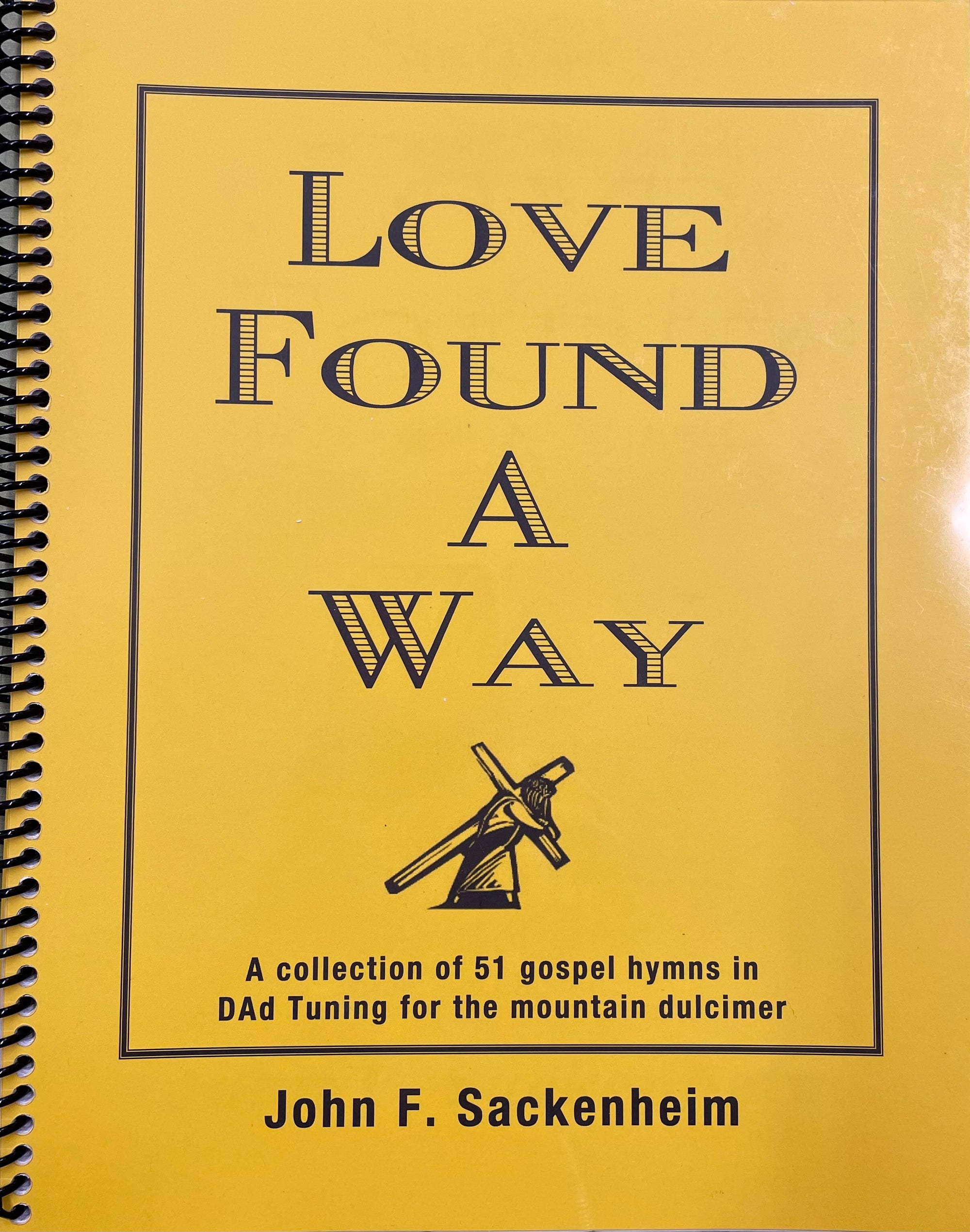 A yellow spiral-bound hymns book titled "Love Found a Way by John Sackenheim", featuring a collection of 51 gospel hymns.