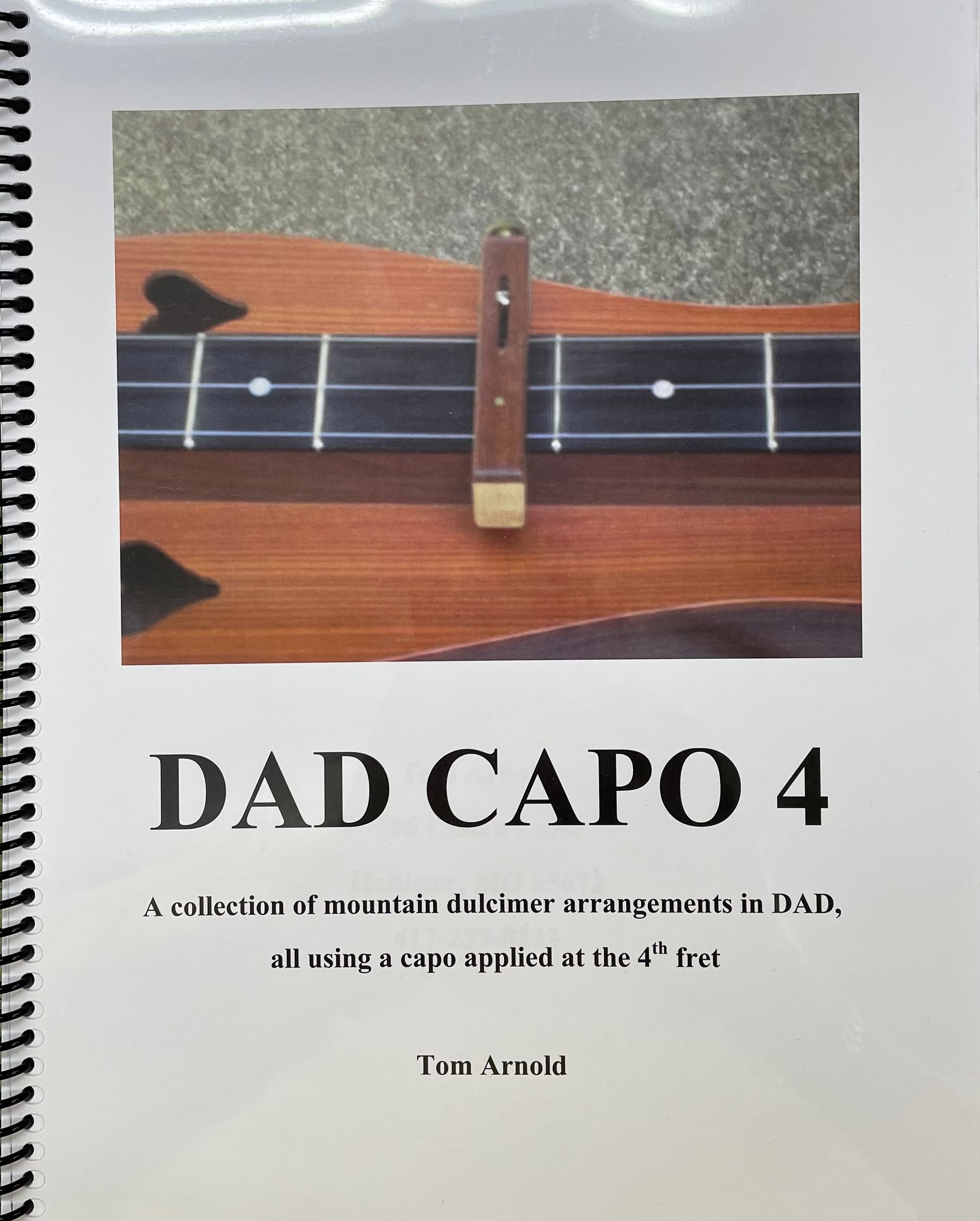 A ukulele with a DAD Capo 4 placed on the fourth fret, tabbed in an old-time tunes collection, featured on the cover of a music book titled "DAD Capo 4" by Tom Arnold.