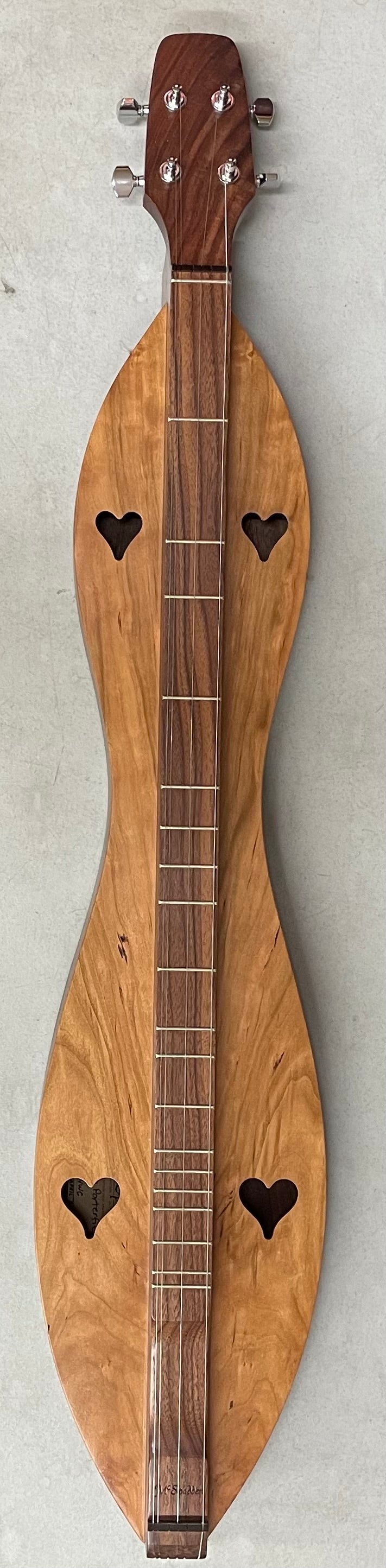 A handcrafted 4 String, Flathead, Hourglass, with Walnut back and Cherry sides and top (4FHWC), featuring heart-shaped sound holes and six tuning pegs. The fretboard is dark wood, contrasting beautifully with the lighter wooden body. Perfect for a beginner dulcimer player, this exquisite instrument rests elegantly on a flat surface.