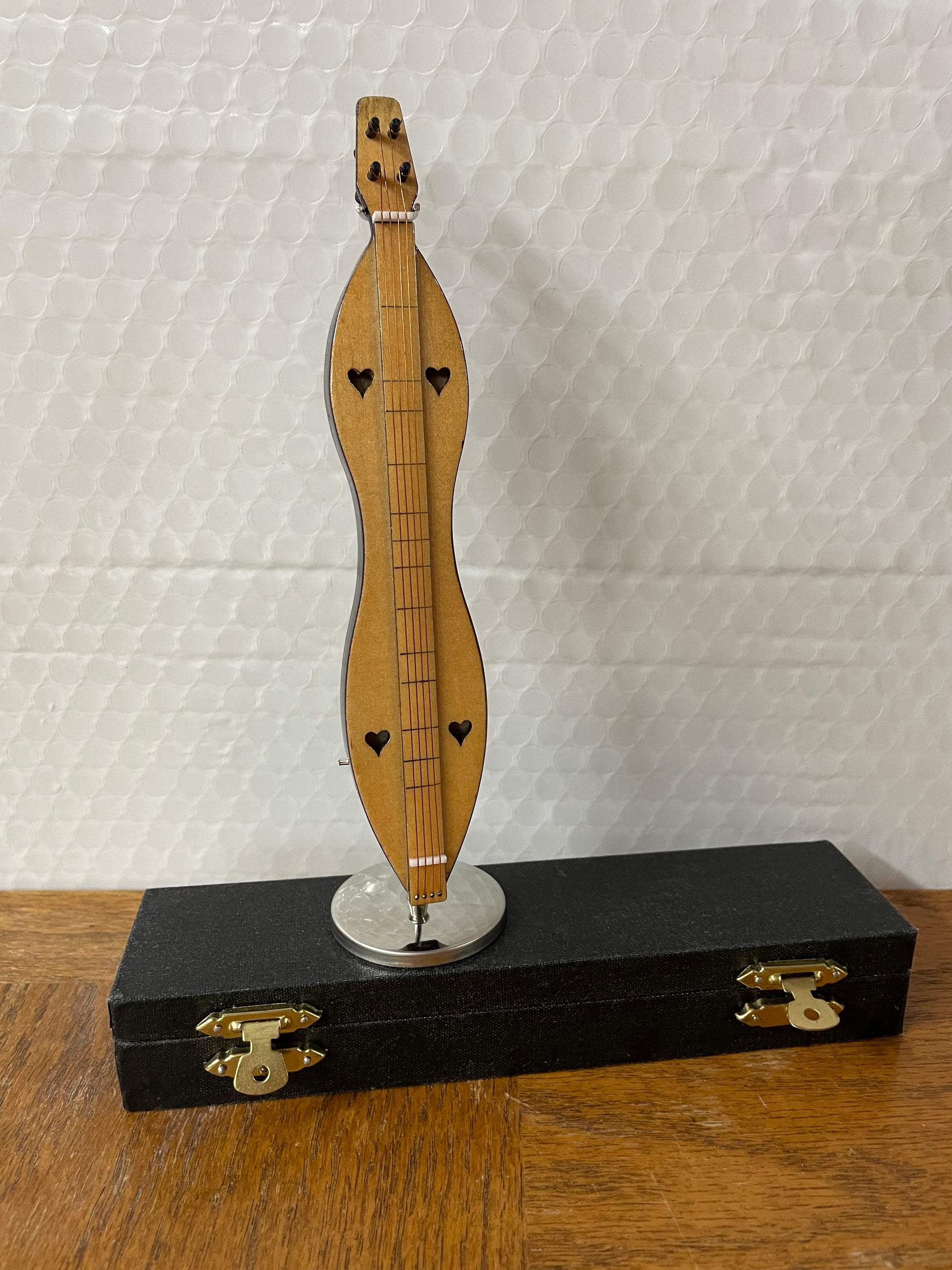 The 7" Dulcimer Miniature with heart-shaped sound holes is displayed upright on a black rectangular stand.