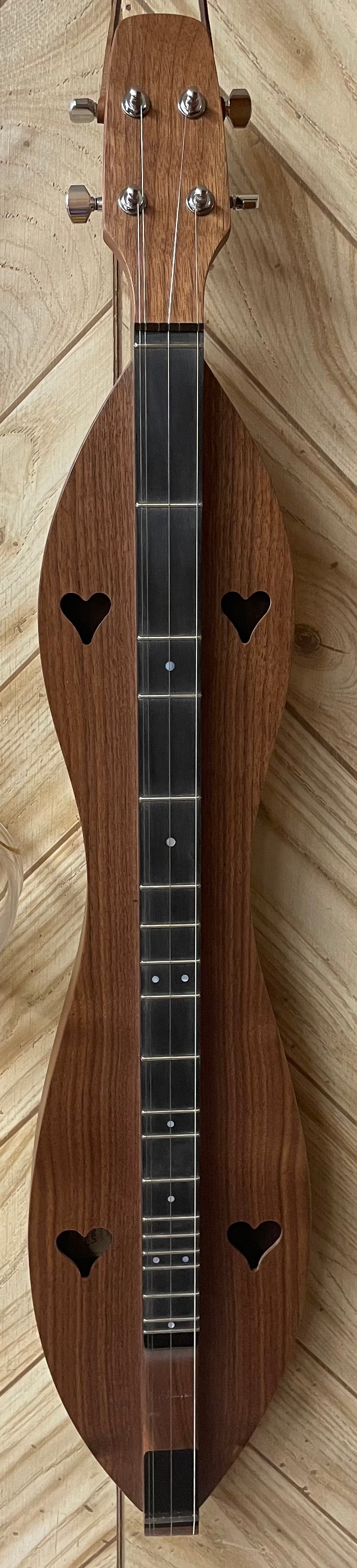 A wooden dulcimer featuring heart-shaped sound holes, an hourglass design, and four tuning pegs, along with a dark chromatic fretboard highlighted by Mother of Pearl Inlay Fret Dots, is resting on a wooden surface.