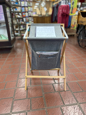 A wooden and fabric *Sling Music Stand* elegantly stands on the tiled floor of a store, showcasing magazines in its main compartment and pocket. This handcrafted stand adds a touch of charm, inviting those on the waiting list for exclusive handmade items to explore further.