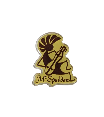 Yellow and brown McSpadden Kokopelli Pin depicting a stylized figure playing a mountain dulcimer, with the word “McSpadden” written below the illustration.