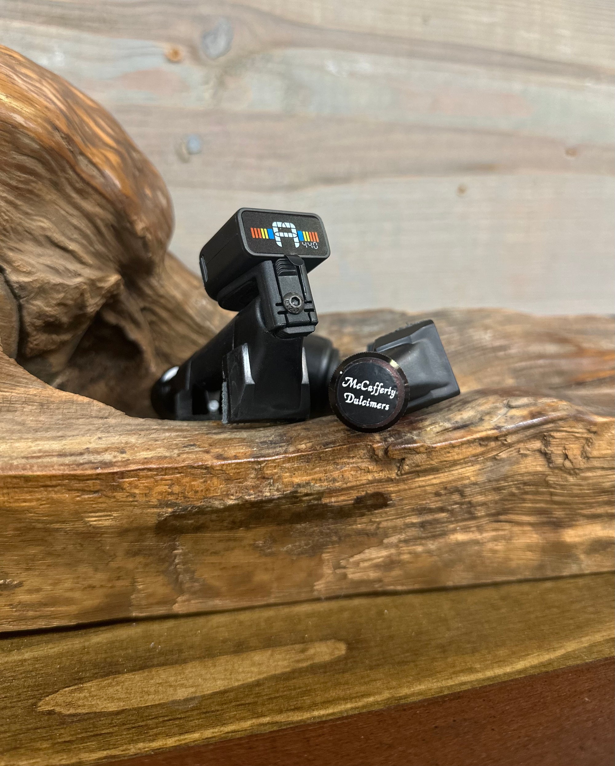 Two robust McCafferty Capo With Tuner devices sit on a wooden surface, one turned over to display the brand name. The textured wood panel backdrop enhances the sleek design of these six-string accessories, complete with flexible silicone grips.