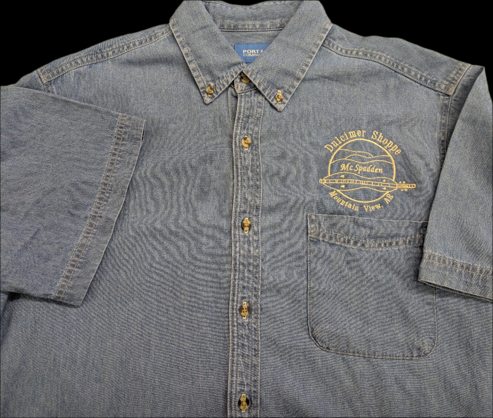 The Denim Short Sleeve Shirt features a gray denim design with the Dulcimer Shoppe logo and dulcimer graphic embroidered above the front pocket.