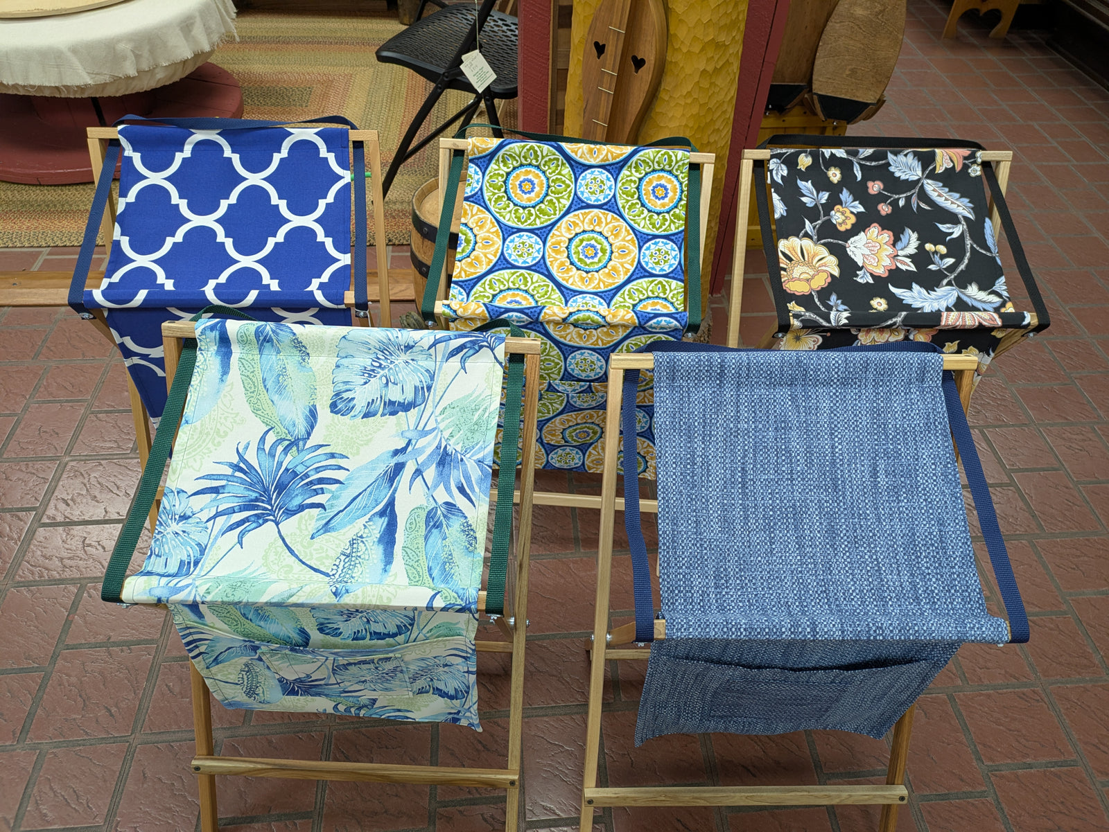 Five folding canvas chairs with wooden frames, each in a different patterned fabric, are arranged on an indoor red tile floor next to a handmade Sling Music Stand.