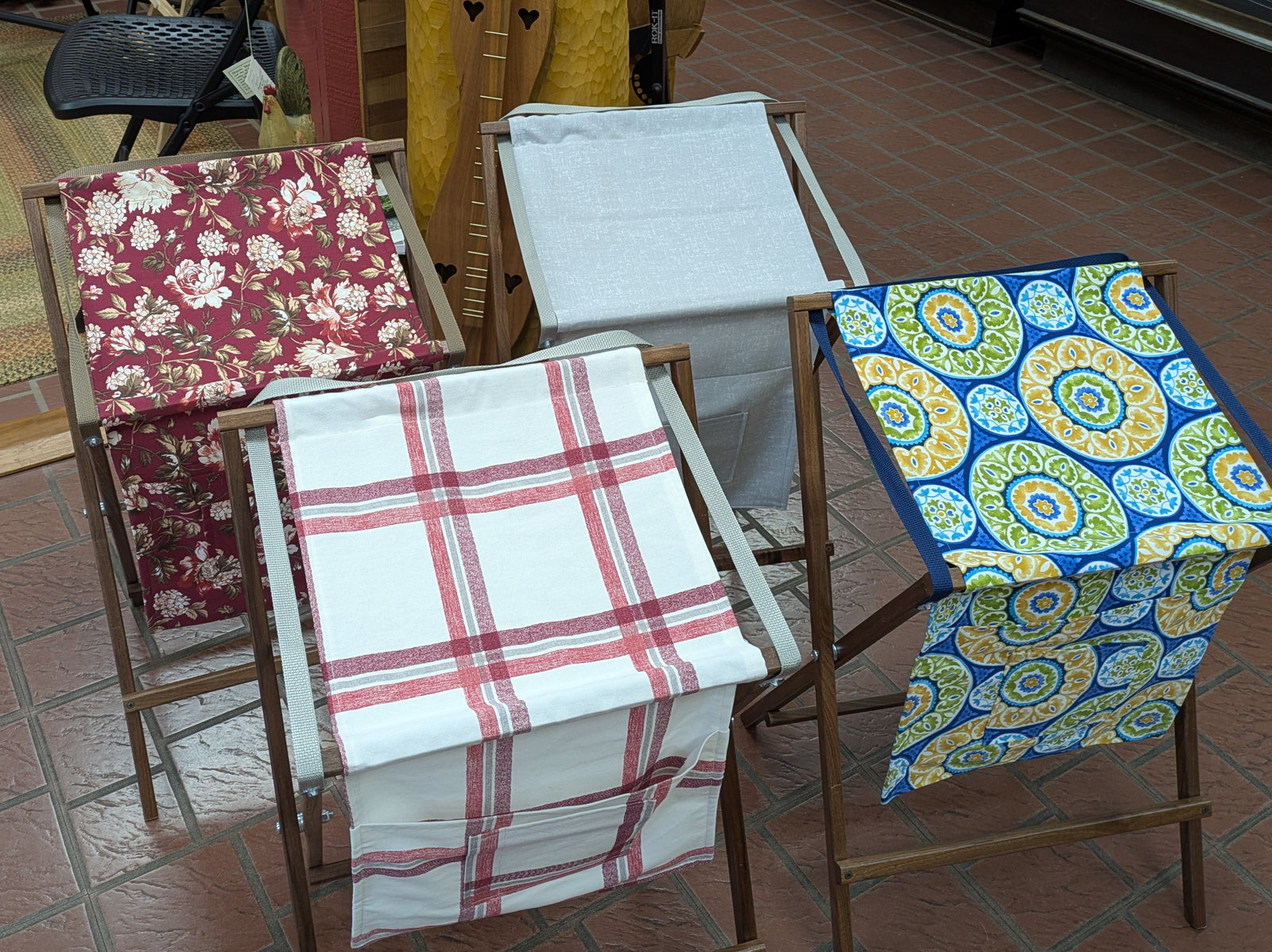 Four Sling Music Stands, reminiscent of folding wooden racks or instrument accessories, display cloth towels with different patterns—red floral, plain white, red plaid, and blue-yellow geometric—on a brown tiled floor indoors.
