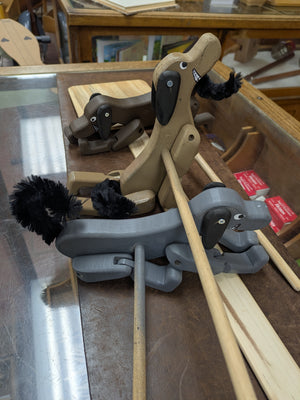 Three Dancing Dog wooden pull toys, handcrafted in Texas and finished with black yarn tails, are displayed on a glass counter. Each Dancing Dog is attached to a wooden stick for pushing—making them perfect for kids’ entertainment or sparking imaginative dancing dog adventures of your own.