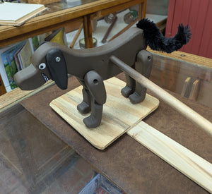 Handcrafted in Texas, the Dancing Dog features movable legs and a stick tail, making it an entertaining wooden toy for kids. It is mounted on a rectangular wooden base and displayed on a glass counter with shelves in the background.