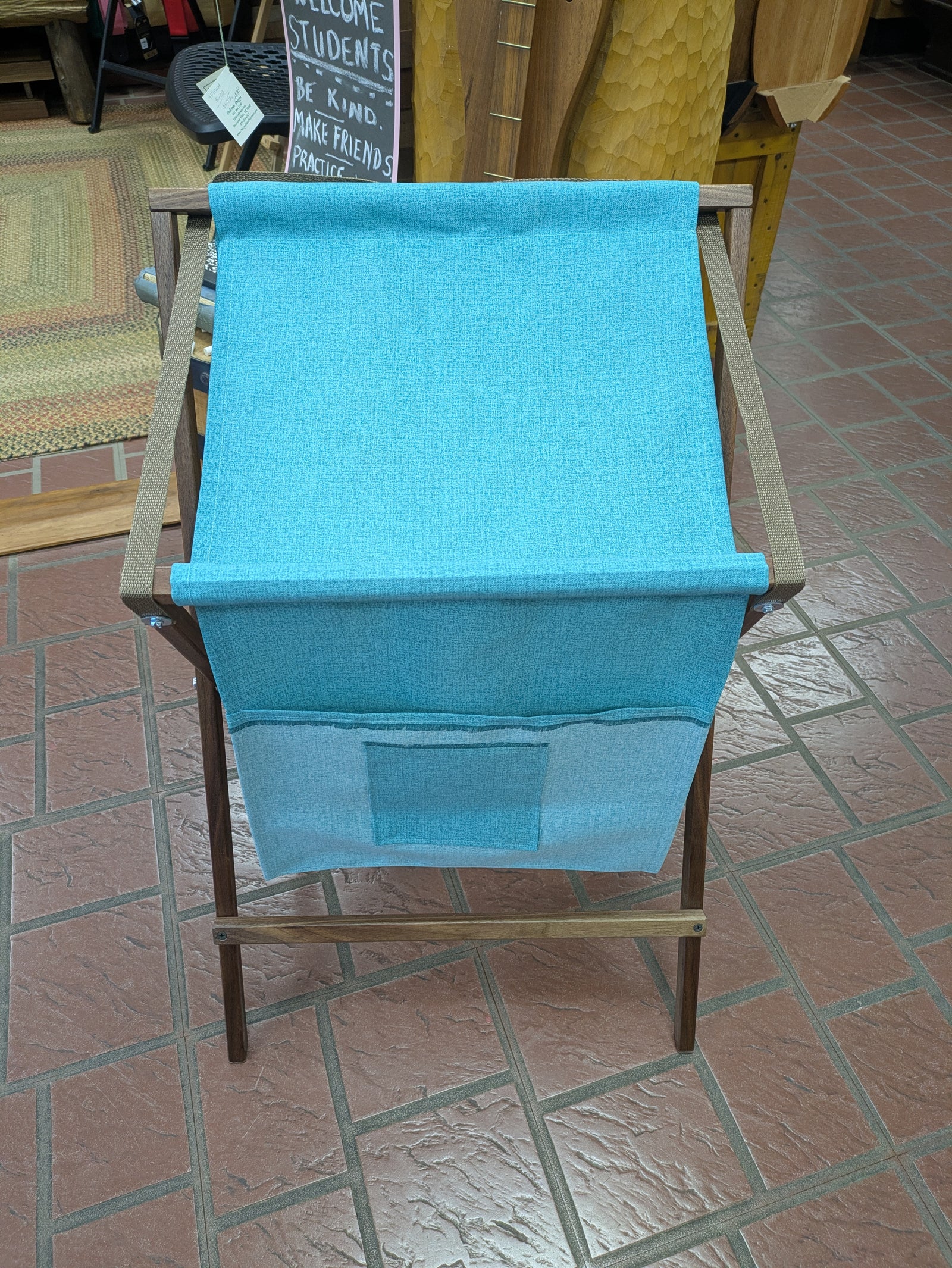 The Sling Music Stand features a wooden frame and a blue fabric seat with a convenient small front pocket, set on an indoor tiled floor. This handmade stand is ideal for musicians, serving both as an instrument accessory or a compact music stand.