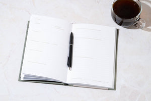 My Travel Journal, featuring lined pages, comes with a black pen and is shown open on a marble surface beside a clear glass cup of black coffee—ideal for capturing your travel memories.