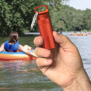 A hand holding a set of two Trek Tubes, waterproof aluminum containers, with carabiners, over a backdrop of a person kayaking on a river, showcasing essential outdoor adventure gear.