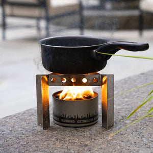 A small Portable Camp Stove / Stand Combo with a lit flame heating a black cooking pot outdoors, placed on concrete near a bench.