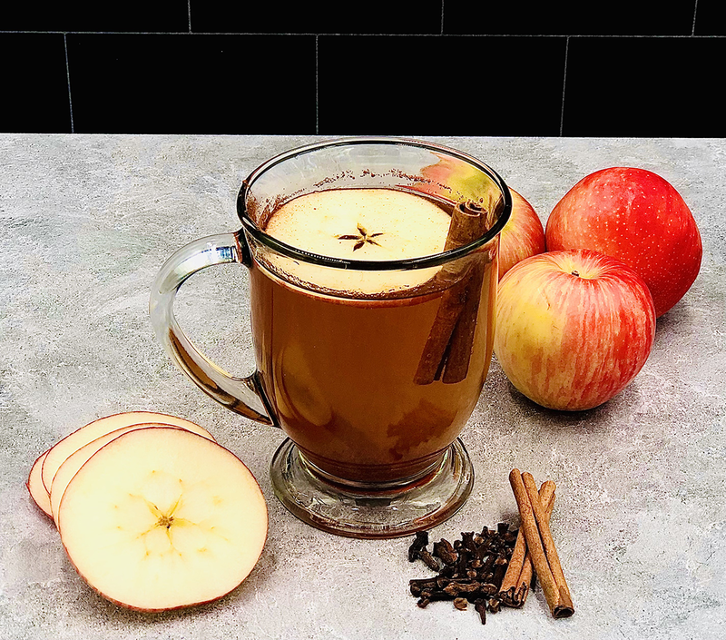 A glass mug filled with apple cider garnished with an apple slice and a Saigon cinnamon stick, surrounded by whole apples, apple slices, and a small pile of cinnamon sticks and cloves on the table, featuring our exquisite Mulling Spices. This festive beverage addition makes for a perfect holiday gift.