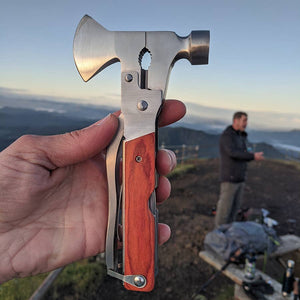 A hand holds an Axe Multi-Tool with a bottle opener against a landscape backdrop at sunset, with a blurred figure standing in the background.