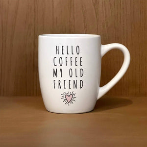 A "Hello coffee, my old Friend" white coffee mug, featuring the text "HELLO COFFEE MY OLD FRIEND" with a small heart icon beneath it, is set against a wooden background next to cozy towels.