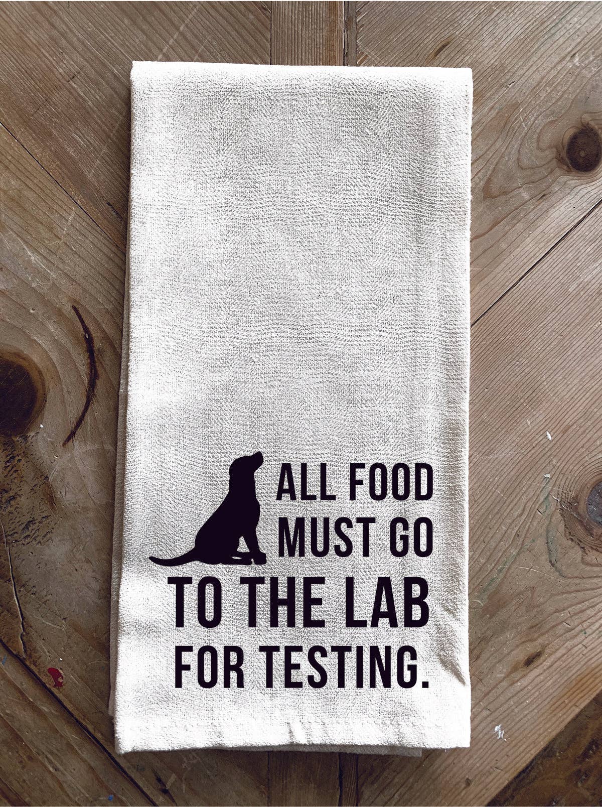 A 100% cotton "All Food must go to the lab for testing" kitchen towel, featuring the silhouette of a dog, placed on a wooden surface.