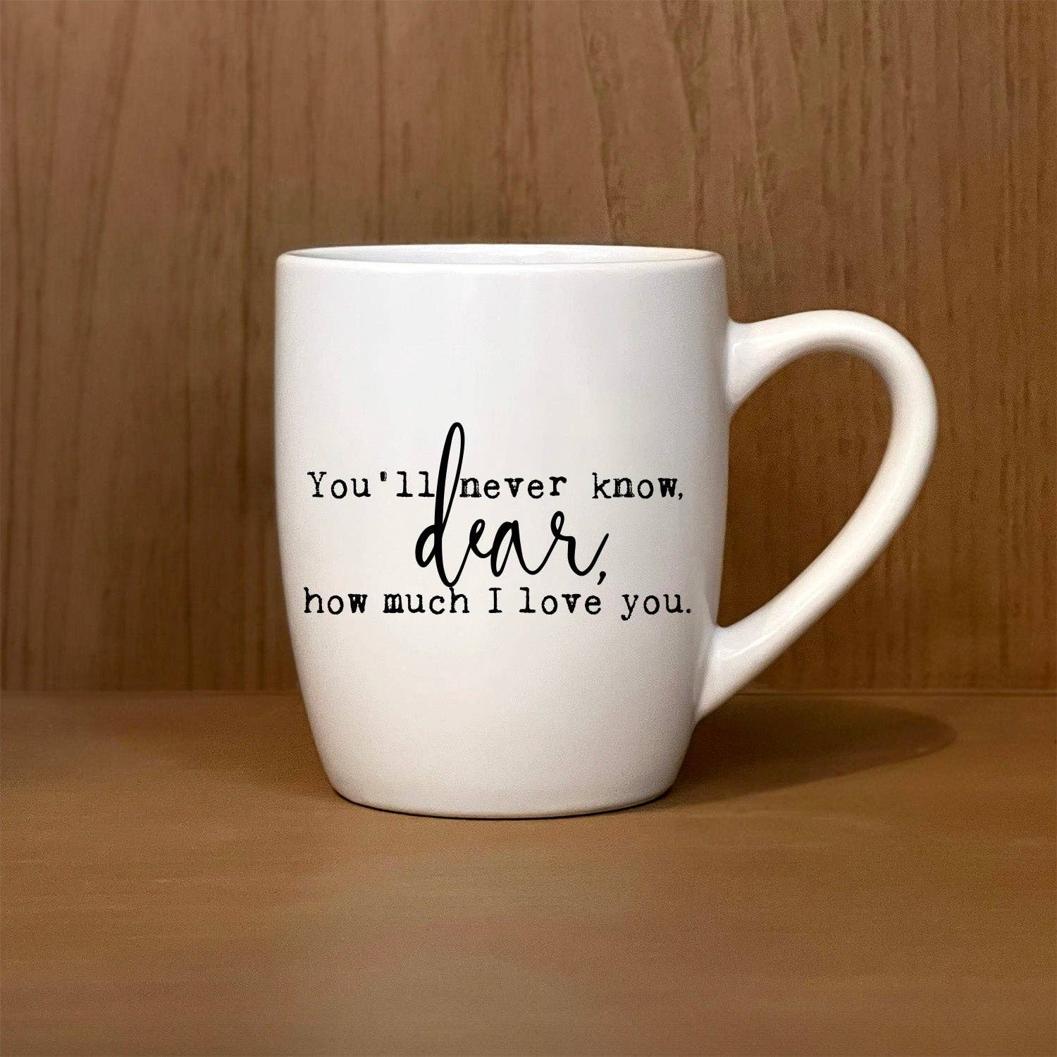 A white mug, labeled "You'll never know, dear, how much I love you," rests on a wooden surface.