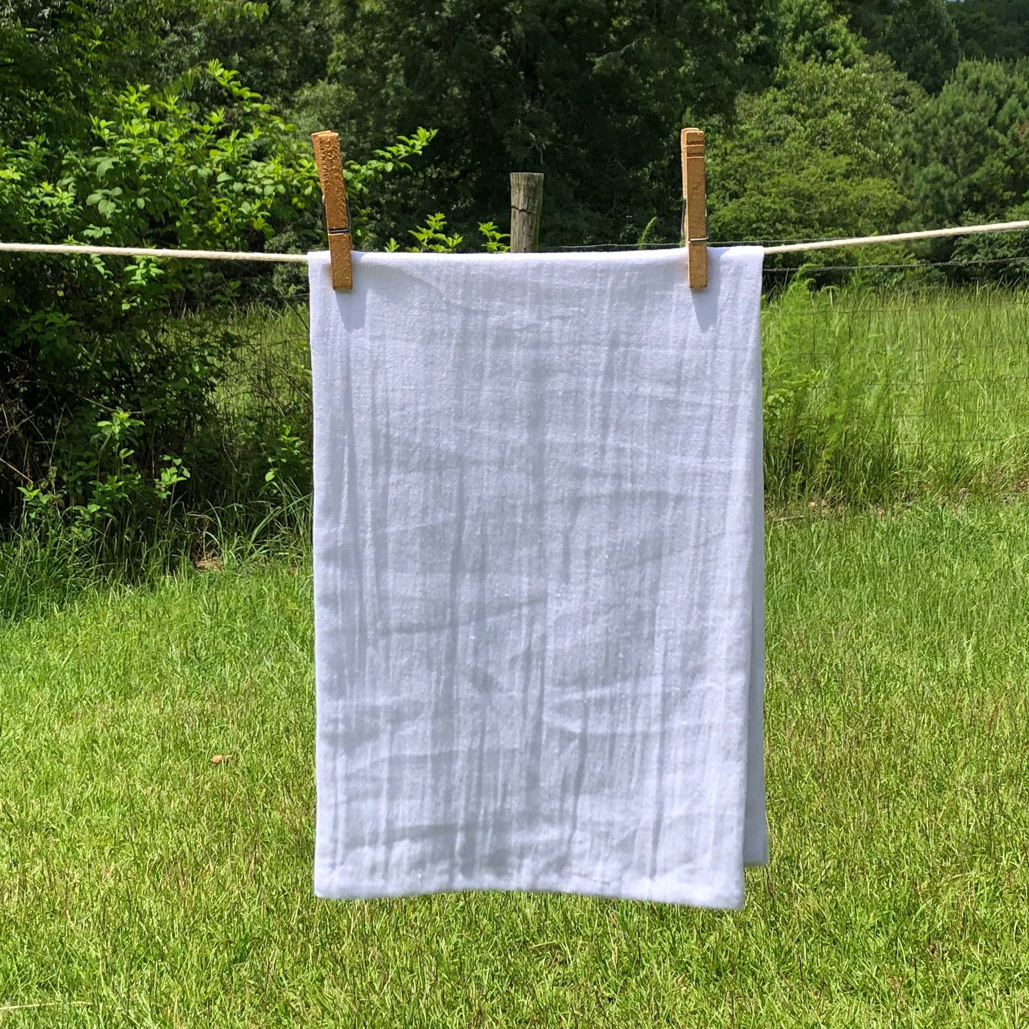 A white Y'all Tea Towel hangs on a clothesline with wooden clothespins in an outdoor, grassy area with green foliage in the background.