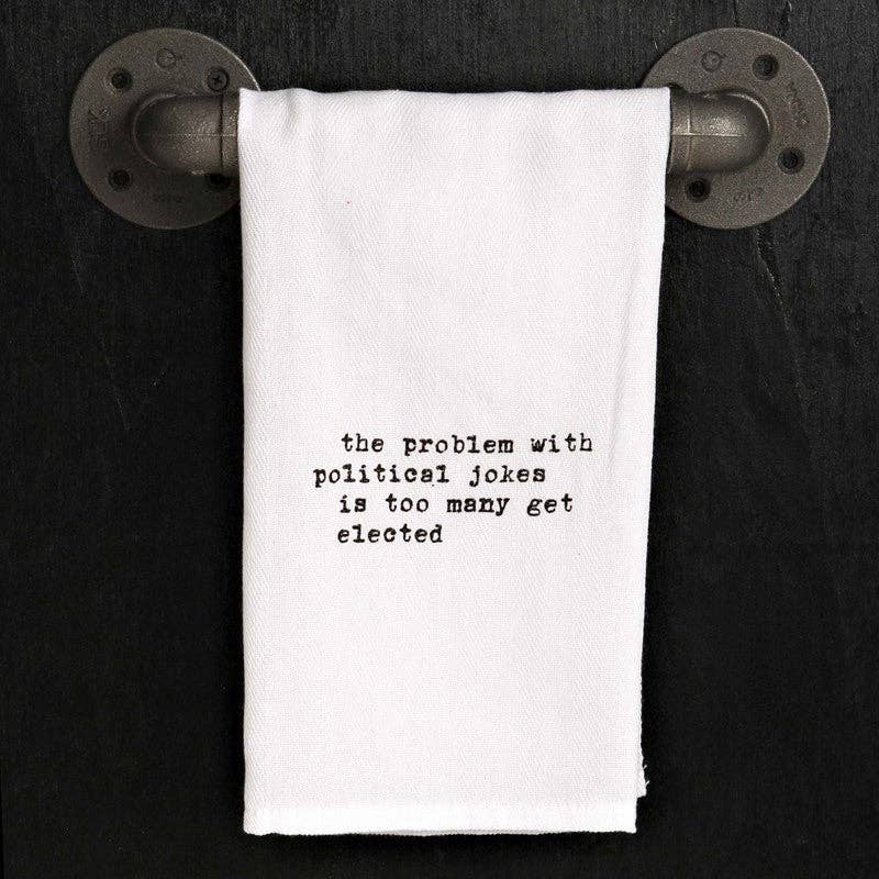 A handmade kitchen towel with black text that reads, "The problem with political jokes is too many of them get elected," hangs on a metal pipe rack against a dark background. This 100% cotton towel, named after its witty message, effortlessly combines humor and functionality, providing both charm and practicality.