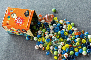 Neato! Marbles In A Tin Box spilled onto a gray fabric surface, showcasing a variety of patterns and various sizes and colors.