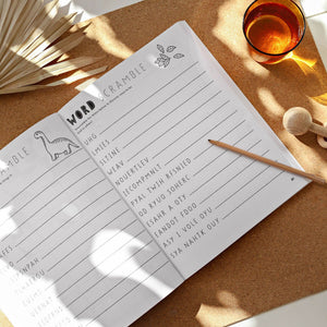 An open copy of "Gratitude For Kids (Mindfulness Activity Book for Kids)" on a corkboard with a pencil, featuring a dinosaur word scramble and a glass of amber liquid—ideal for fun and gratitude as children play and learn.