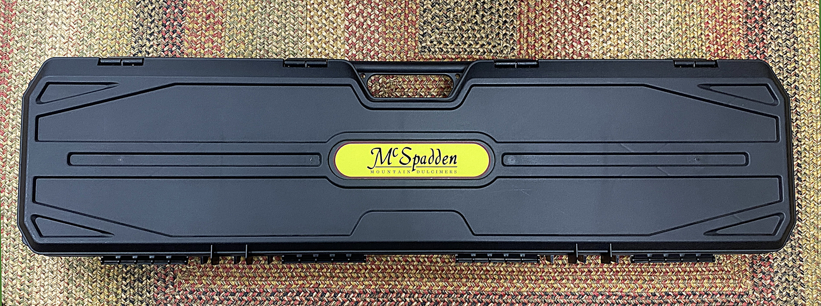 A Hard Shell Dulcimer Case UPGRADE, designed to fit your instrument perfectly, features a black plastic exterior with a textured surface and comes with a yellow and orange "M Spuden Musical Services" label.