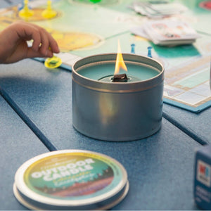 A small, lit portable camping stove on a table with a flame burning, surrounded by various outdoor gear, a map, and eco-friendly candles including The Outdoor Candle - Citronella and Eucalyptus.