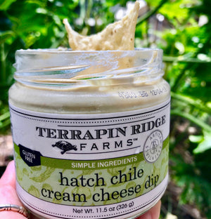 A hand holding a jar of gluten-free Terrapin Ridge Farms Hatch Chile Cream Cheese Dip with a tortilla chip dipped into it, against a backdrop of green foliage.