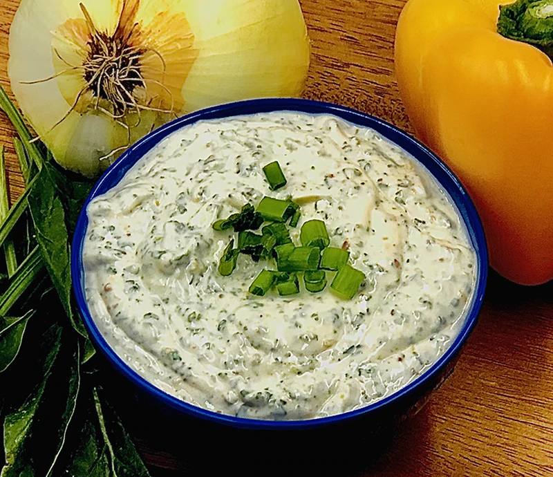 A bowl of Fiesta Spinach Dip Mix garnished with chopped green onions, surrounded by a yellow bell pepper, onion, and fresh spinach leaves on a wooden surface.