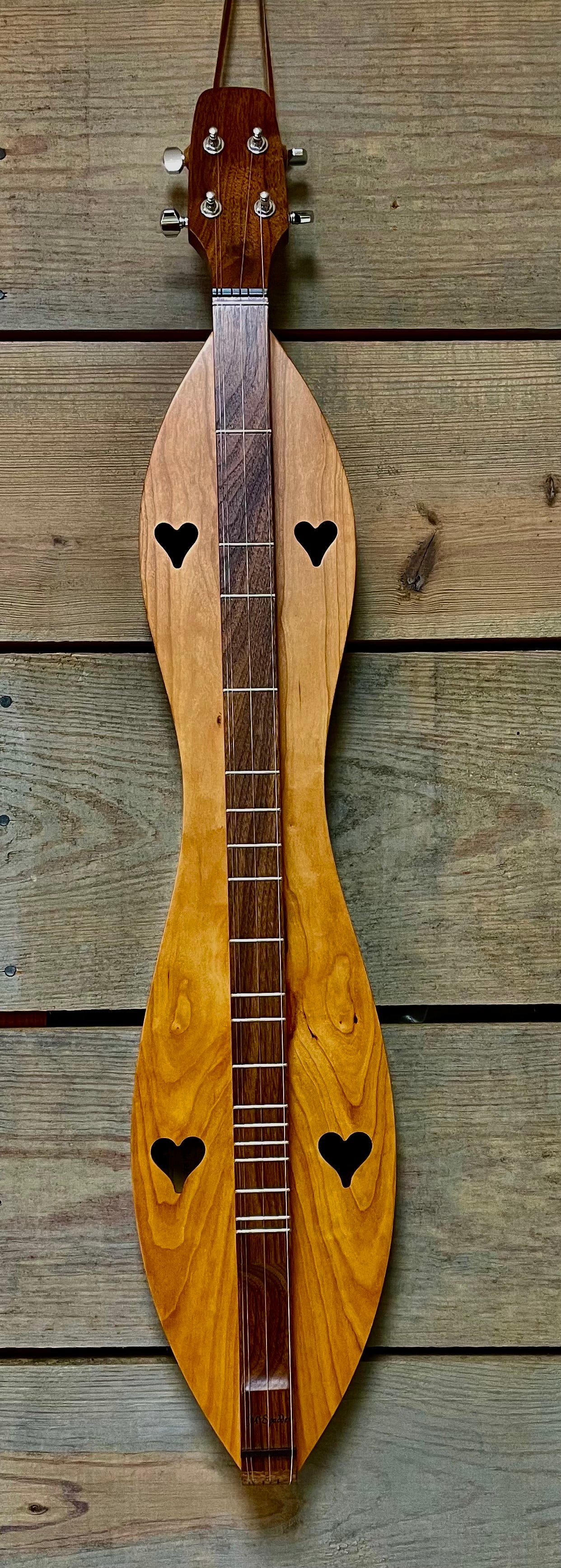 This image shows a 4 String, Flathead, Hourglass dulcimer with a Cherry back and Walnut sides and top (4FHCW), a handcrafted wooden stringed instrument featuring heart-shaped sound holes and a narrow, elongated body, hanging on a wooden wall.