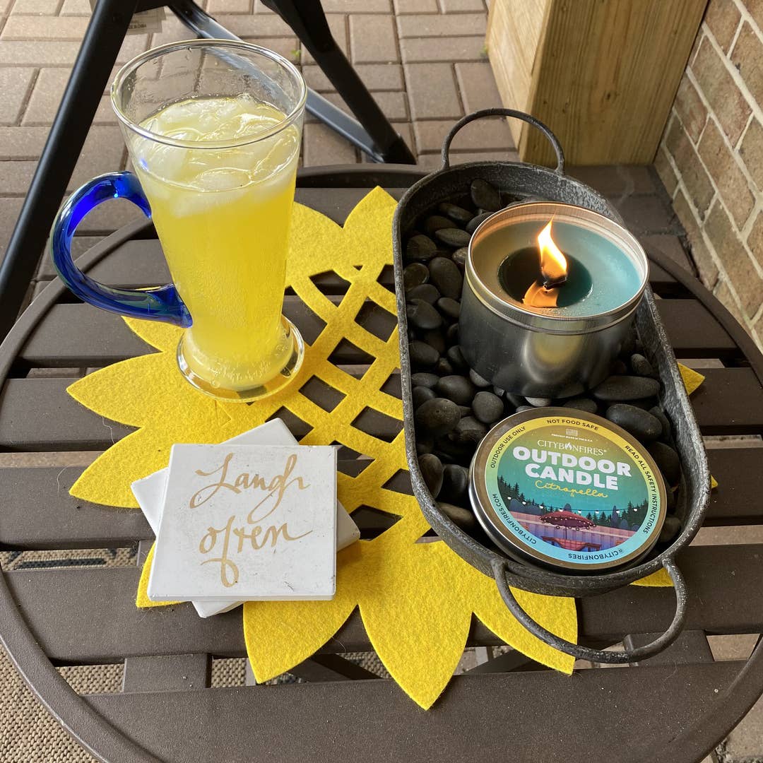 A refreshing drink in a glass beside an Outdoor Candle - Citronella and Eucalyptus in a tin, set on a black tray with rocks, next to a "laugh often" sign on a yellow star-shaped mat