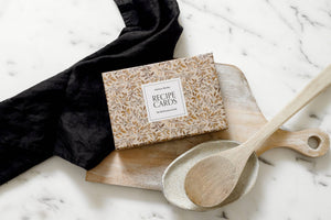The Recipe Cards 4x6 sit on a wooden cutting board with ceramic and wooden spoons and a folded black cloth on a marble countertop—an ideal kitchen or hostess gift.