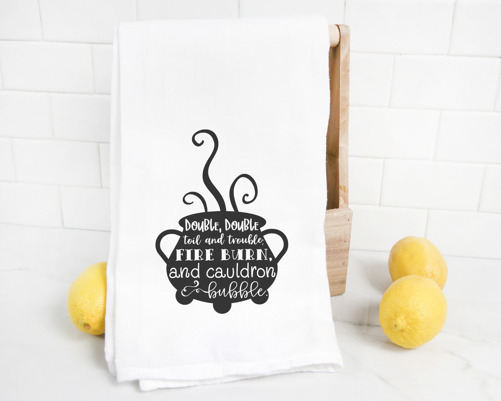 The discontinued “Double Double Toil and Trouble” tea towel is a cotton tri-fold towel with a black cauldron and Shakespeare’s quote—an ideal whimsical Halloween kitchen accent. Shown with three lemons and a wooden box.